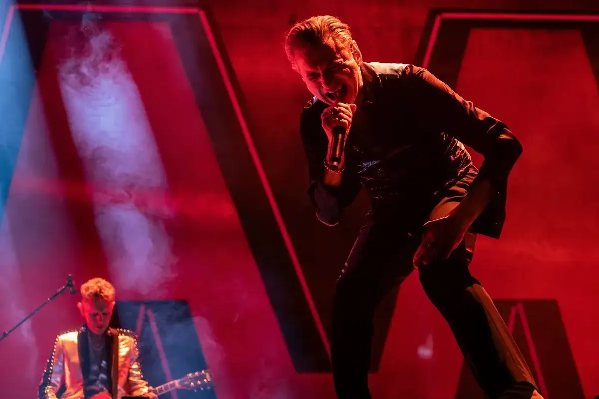 Dave Gahan śpiewa na koncercie Depeche Mode w Warszawie. W tle widać gitarzystę i czerwone światła.
