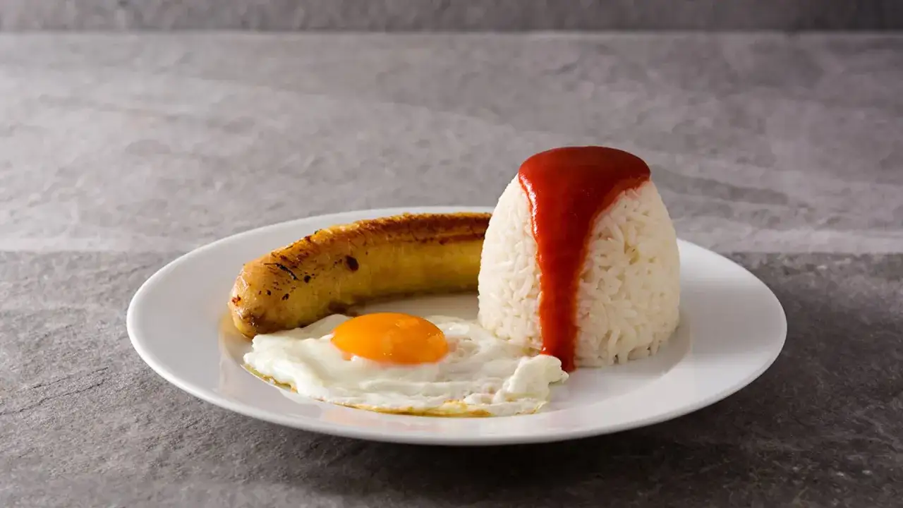 Arroz a la cubana: arroz blanco con salsa roja, huevo frito y pl&aacute;tano maduro frito en un plato blanco.