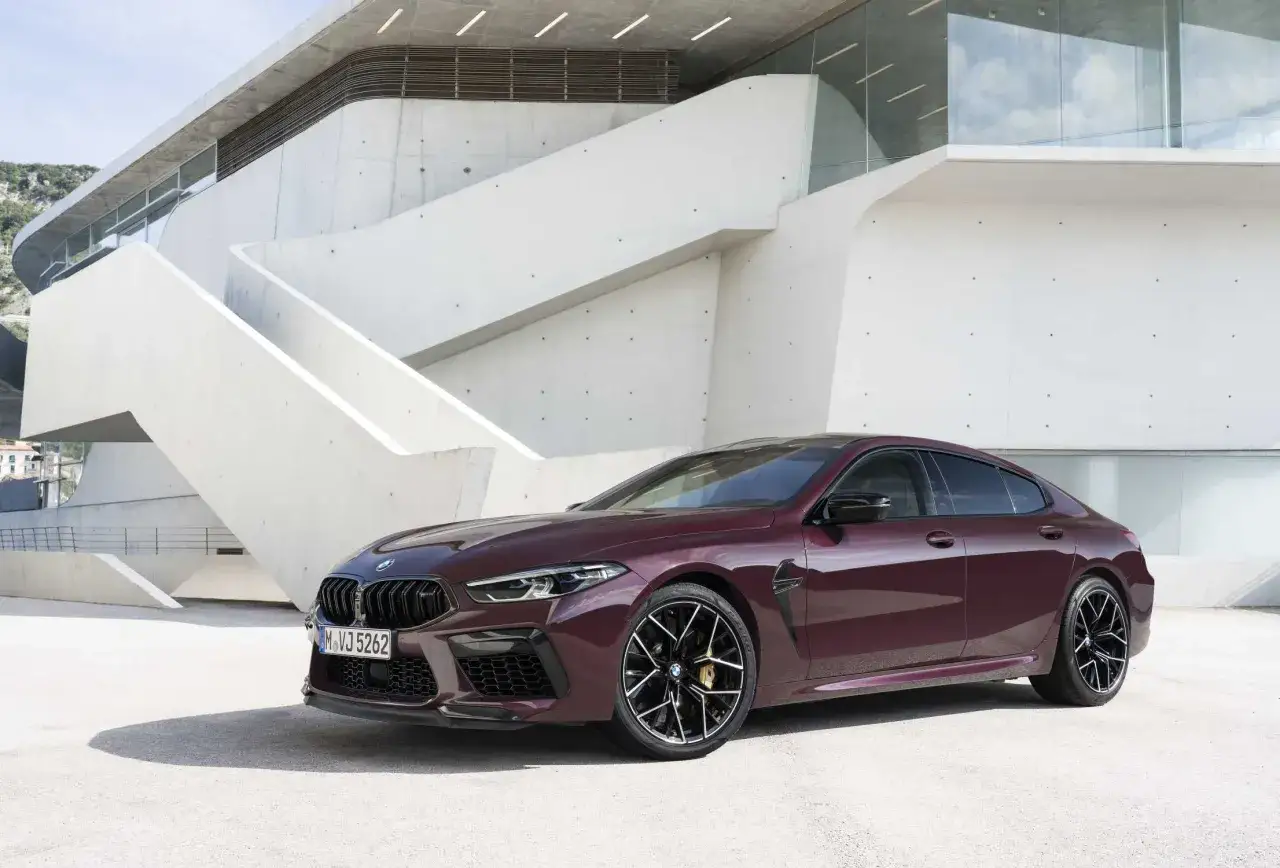 Eleganckie, bordowe BMW M8 Gran Coupe zaparkowane przed nowoczesnym budynkiem o geometrycznej architekturze.