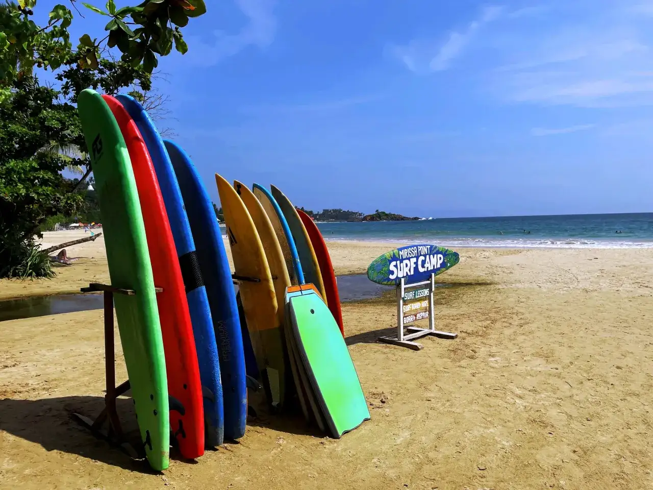 Kolorowe deski surfingowe stoją na plaży w Mirissa Sri Lanka, gotowe na fale. Obok tablica informuje o lekcjach surfingu.