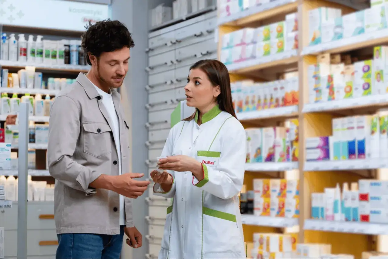 Klient rozmawia z młodą panią technik farmacji w aptece. Ona pokazuje mu małe opakowanie.