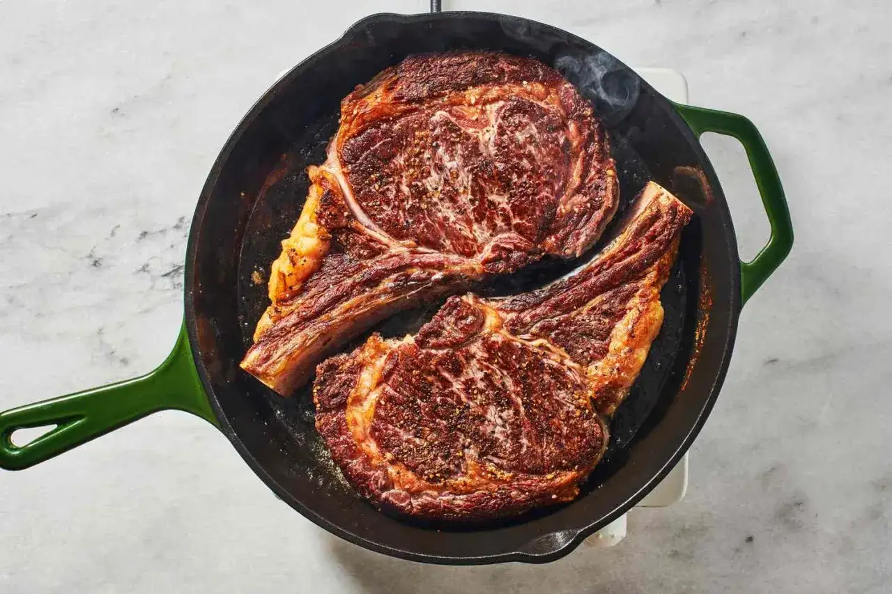 Zwei saftige Rib-Eye-Steaks braten in einer Gusseisenpfanne. Rauch steigt auf, w&auml;hrend sie perfekt gegart werden.
