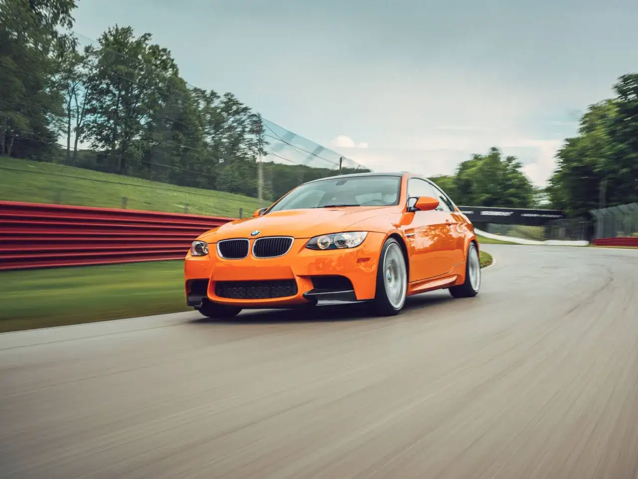 Pomarańczowe BMW M3 E92, zaprojektowane przez Chrisa Bangle'a, śmiga po torze wyścigowym, otoczone zielenią i czerwonymi barierami.