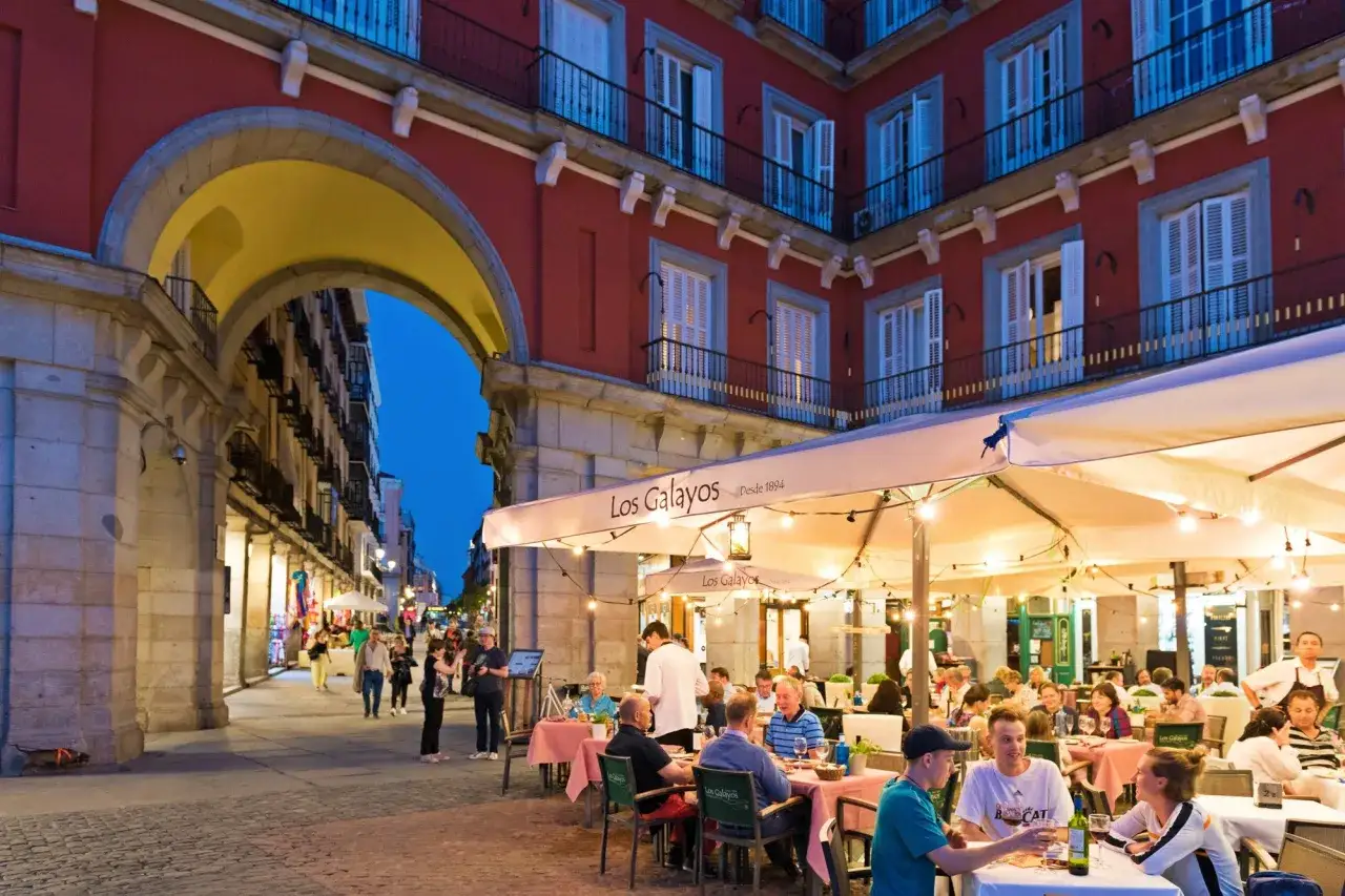 Terrasse anim&eacute;e de Los Galayos, un endroit id&eacute;al pour manger &agrave; Madrid. Des gens d&icirc;nent sous des parasols &eacute;clair&eacute;s par des guirlandes lumineuses.