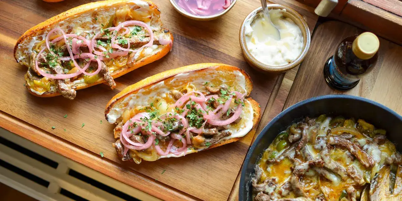 Tv&aring; philly cheese steak-mackor med m&ouml;ra strimlor av k&ouml;tt, sm&auml;lt ost, l&ouml;k och paprika, toppade med picklad r&ouml;dl&ouml;k och persilja.