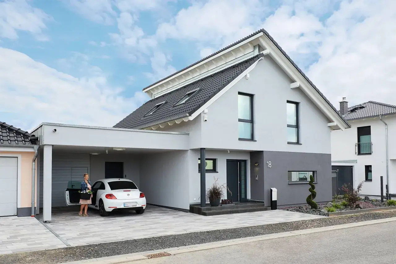 Modernes Haus mit Carport und versetztem Pultdach. Eine Frau steigt in ihren weißen Käfer.