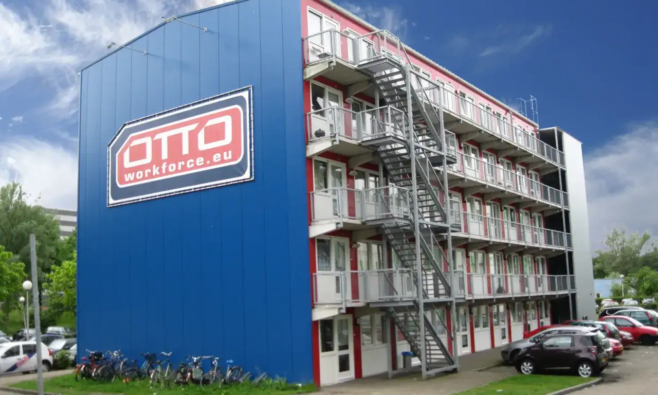 Budynek z niebieskim panelem i logo OTTO Workforce.eu, oferujący eb personeelsdiensten. Wiele rower&oacute;w i samochod&oacute;w na parkingu.