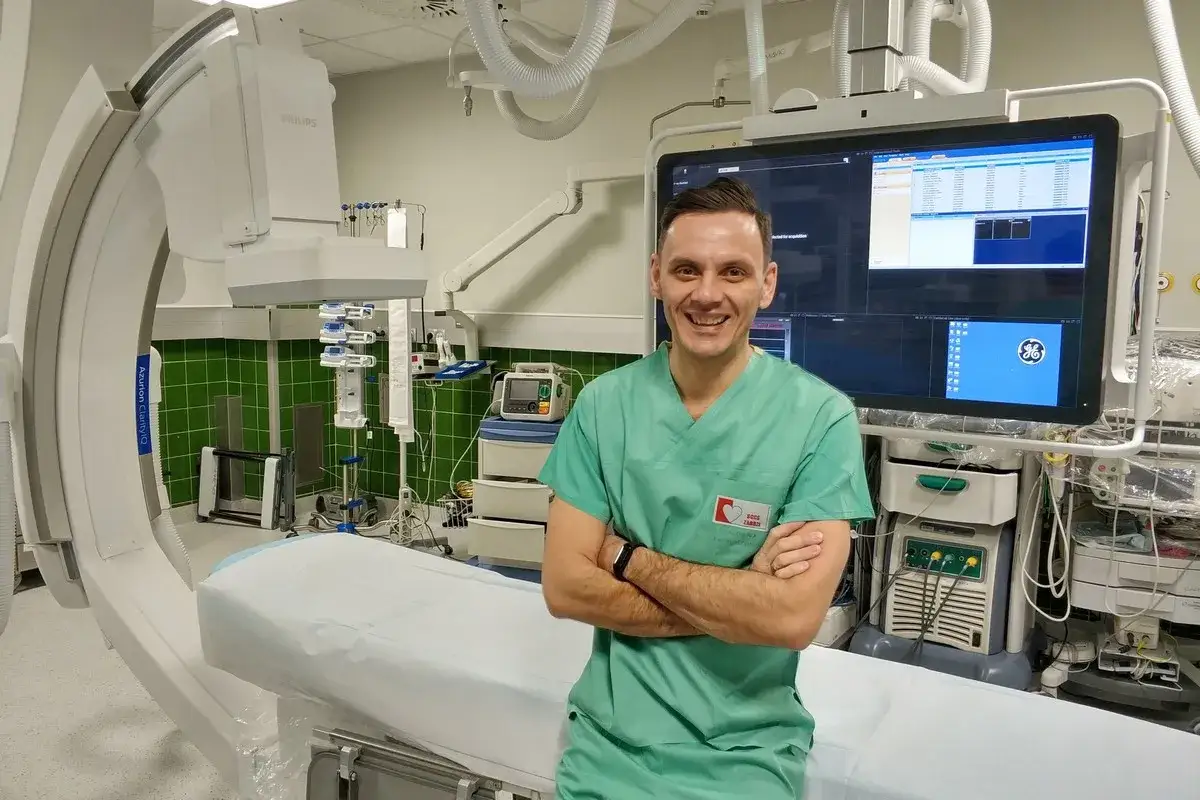 Uśmiechnięty technik elektroradiolog w zielonym fartuchu stoi w sali zabiegowej obok aparatury medycznej.