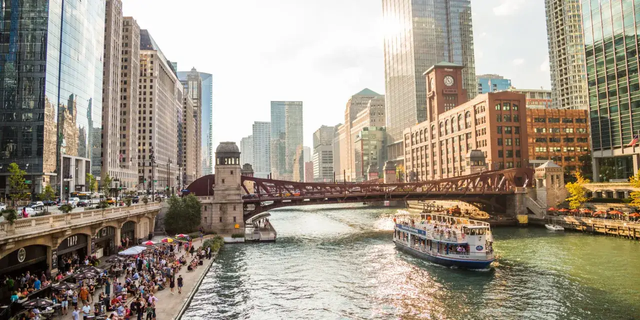 A perfect day in Chicago: a boat tour on the river, people dining al fresco, and iconic architecture under a bright sun.