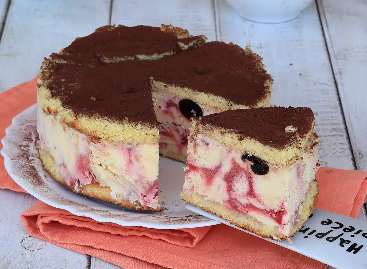 Deliziosa torta gelato con pan di Spagna, crema e ciliegie, spolverata di cacao. Una fetta è già servita su un piattino.