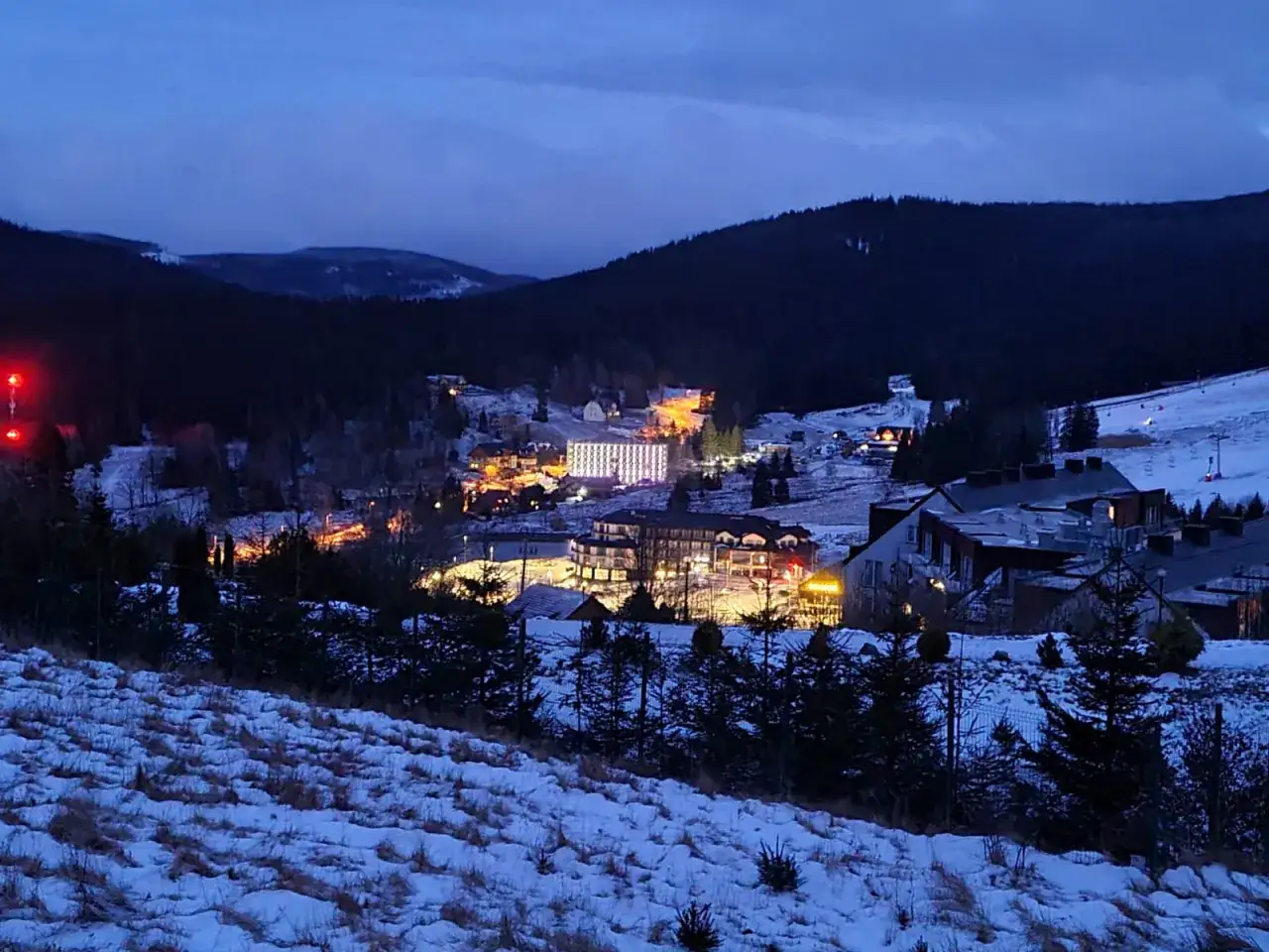 Oświetlony ośrodek górski Czarna Góra Resort w zimowej scenerii, z budynkami i stokami narciarskimi widocznymi w zmierzchu.