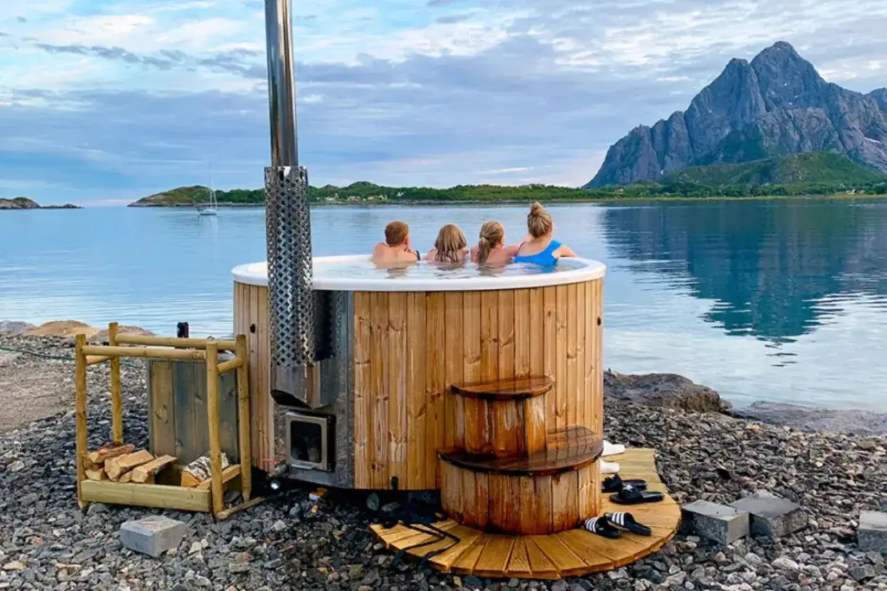 Familj njuter av badtunna vid havet under en ljus lofoten sommar. Berg och segelbåt i bakgrunden.