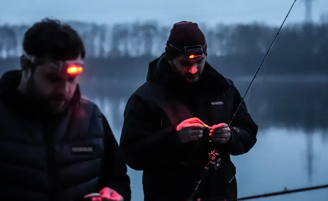 Zwei Angler bereiten sich auf das Nachtangeln vor. Mit Stirnlampen beleuchten sie ihre K&ouml;der und die Angelruten im D&auml;mmerlicht.