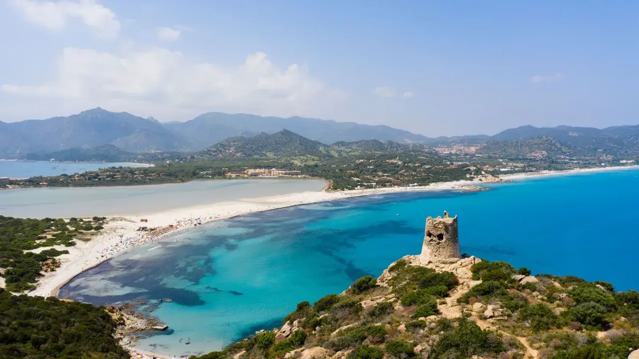 En ruinerad vakttorn vakar över den turkosa bukten vid Costa Smeralda, Sardinien. En lång sandstrand med parasoller och badande sträcker sig längs kusten.