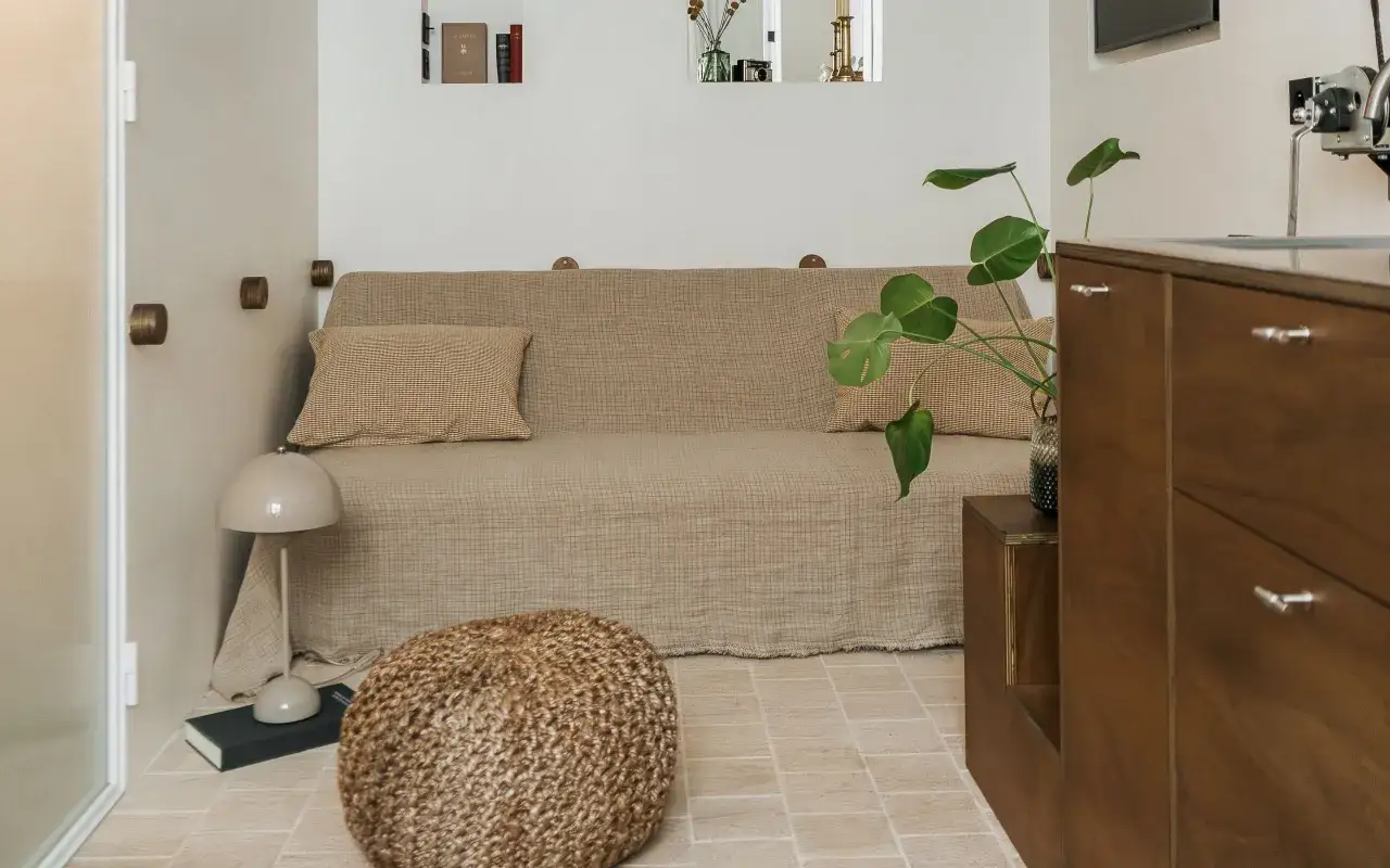 Acogedora habitaci&oacute;n de 4 metros cuadrados con sof&aacute; cama, puf, planta y mueble de madera.