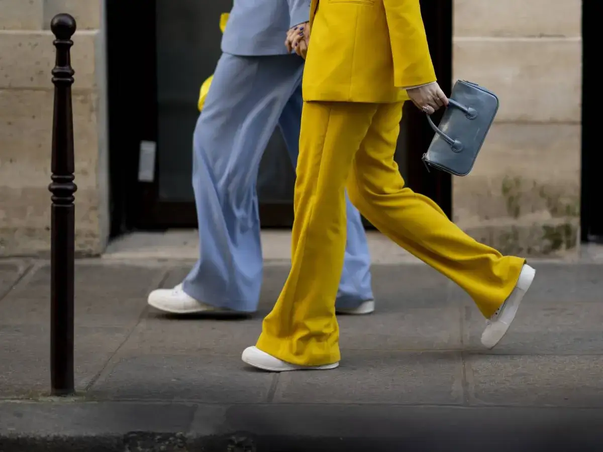 Un outfit tailleur con sneakers, giallo vivace e azzurro pastello, camminano sul marciapiede. Una borsa grigia completa il look.
