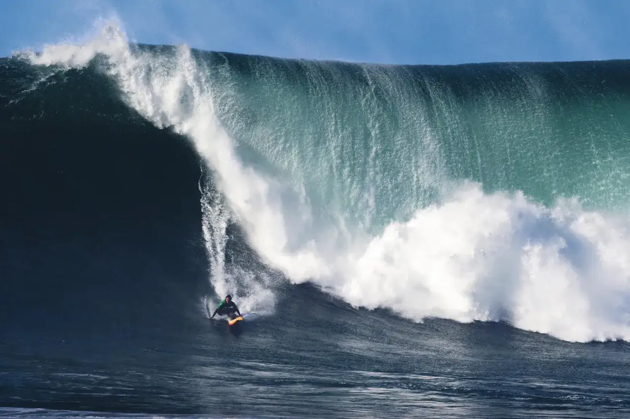 Ein Surfer reitet eine riesige Welle an der Algarve. Die türkisfarbene Gischt bricht über ihm, während er auf seinem Board die Kraft des Ozeans spürt.