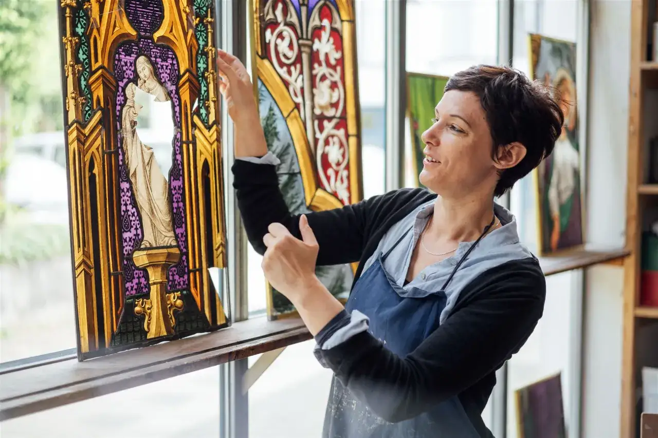 Frau mit Schürze prüft ein buntes Glasfenster. Ihr Studium der Restaurierung zeigt sich in der sorgfältigen Arbeit an historischen Kunstwerken.