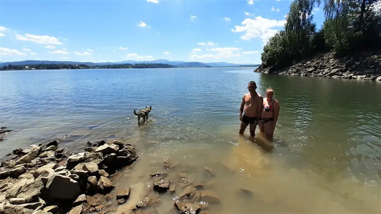 Para z psem na plaży nad Jeziorem Żywieckim. Woda jest płytka, a krajobraz przypomina karaiby.