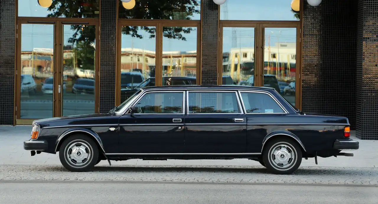 En mörkblå Volvo 264 TE, en lyxig limousin, står parkerad framför en byggnad med stora glaspartier.