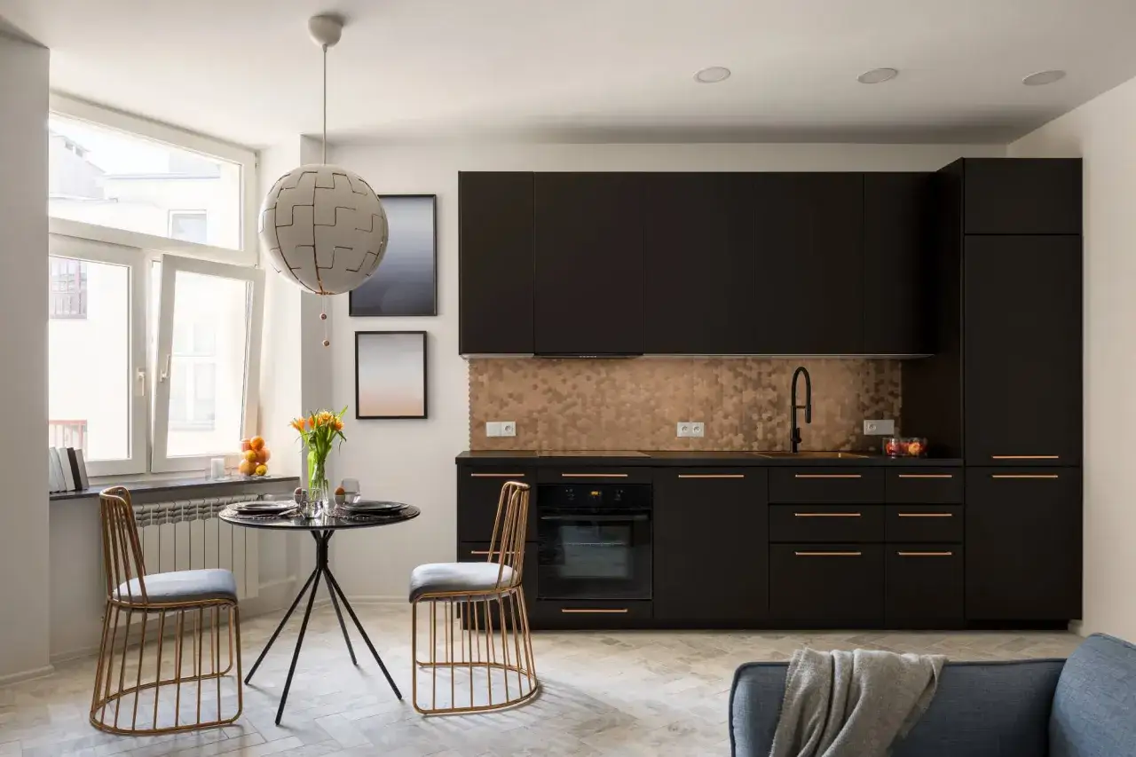 Am&eacute;nagement petite cuisine 2m2 : meubles noirs, cr&eacute;dence hexagonale cuivr&eacute;e, table ronde et chaises dor&eacute;es.