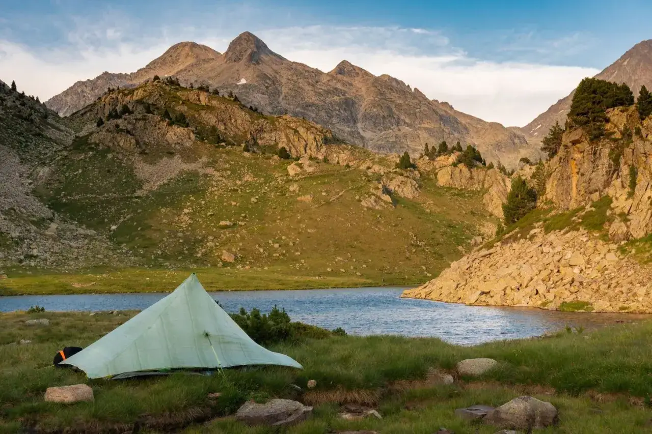 A lone tent sits by a serene lake, a testament to the rugged beauty of the Haute Route Pyrenees. Majestic mountains loom in the background, bathed in the warm glow of sunset.
