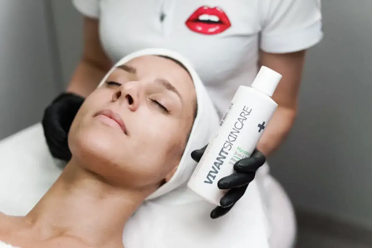 En kvinna med slutna &ouml;gon f&aring;r en ansiktsbehandling. En hand med svart handske h&aring;ller upp en flaska VIVANT SKINCARE, kanske f&ouml;r att behandla oljig hy orsak.