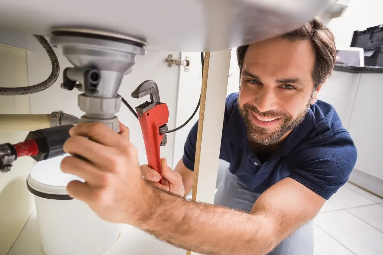 Uśmiechnięty hydraulik naprawia rury pod zlewem, używając czerwonego klucza.