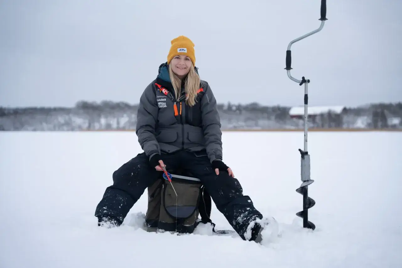 Kvinna ler och njuter av att fiska p&aring; isen. Hon sitter p&aring; en ryggs&auml;ck bredvid en isborr.