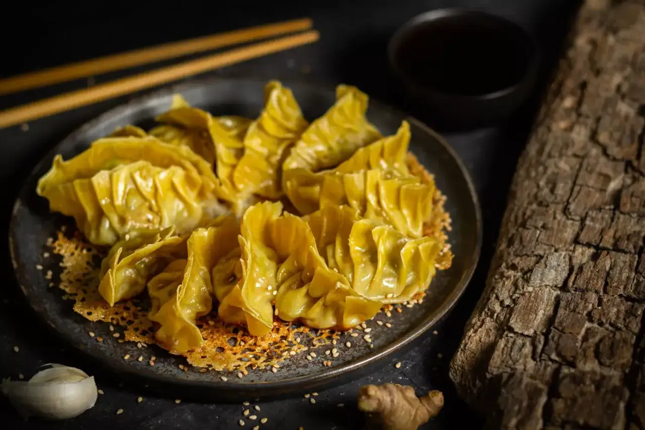 Goldbraune, knusprige Gyoza dumplings auf einem Teller, garniert mit Sesam. Daneben Ingwer, Knoblauch und ein Sch&auml;lchen Sojasauce.