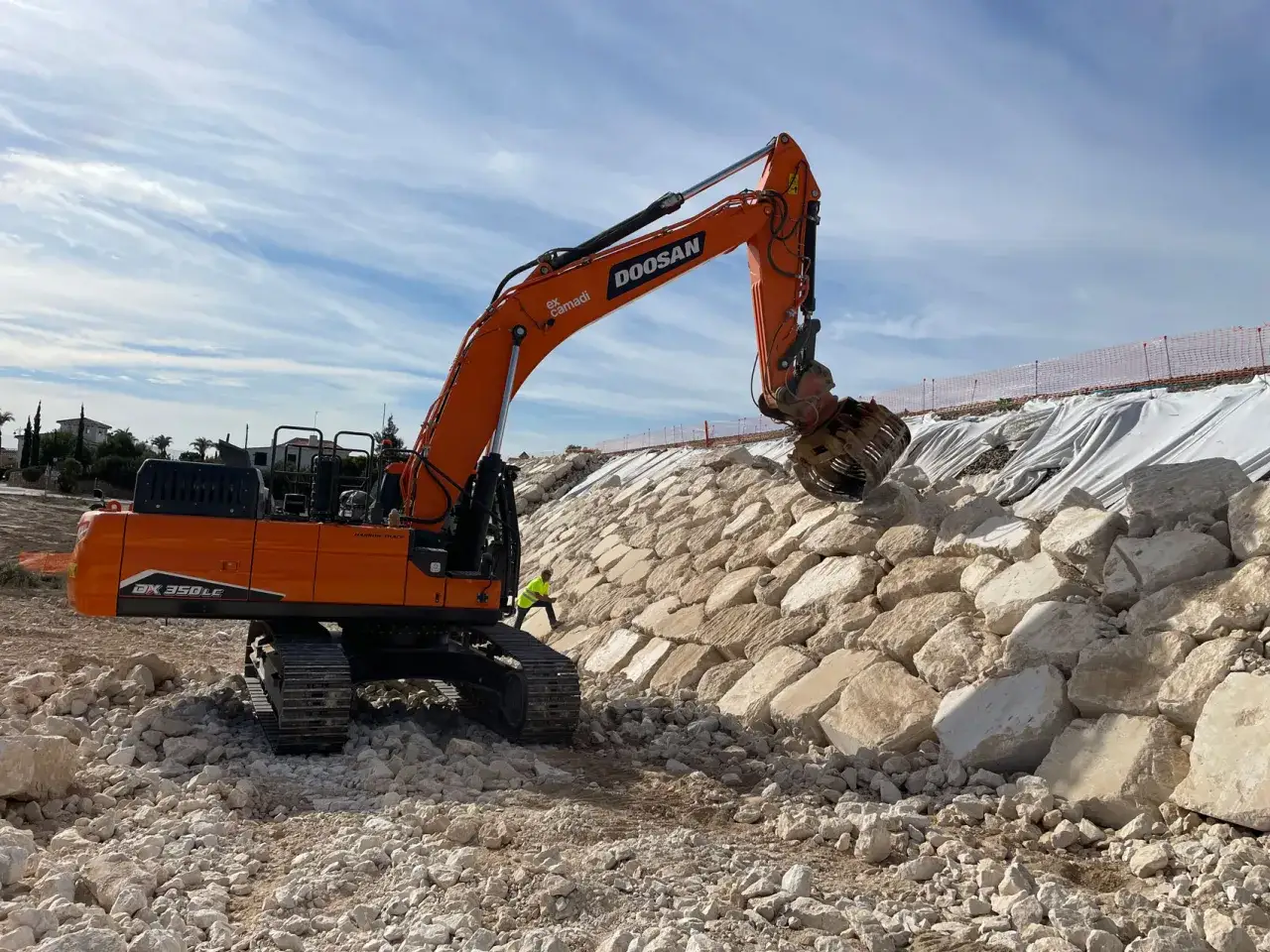 Excavadora naranja trabajando en una escollera, colocando rocas para estabilizar la ladera. El escollera significado es protección costera.