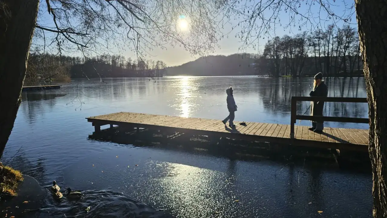 Dzieci cieszą się feriami lubuskimi, spacerując po drewnianym pomoście nad zamarzniętym jeziorem. Słońce odbija się od tafli wody.