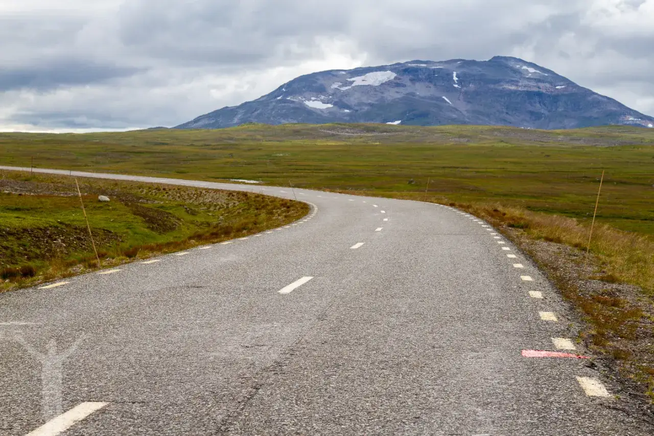 En slingrande väg leder mot ett snöklätt berg i ett vidsträckt landskap. Perfekt för en roadtrip Sverige.