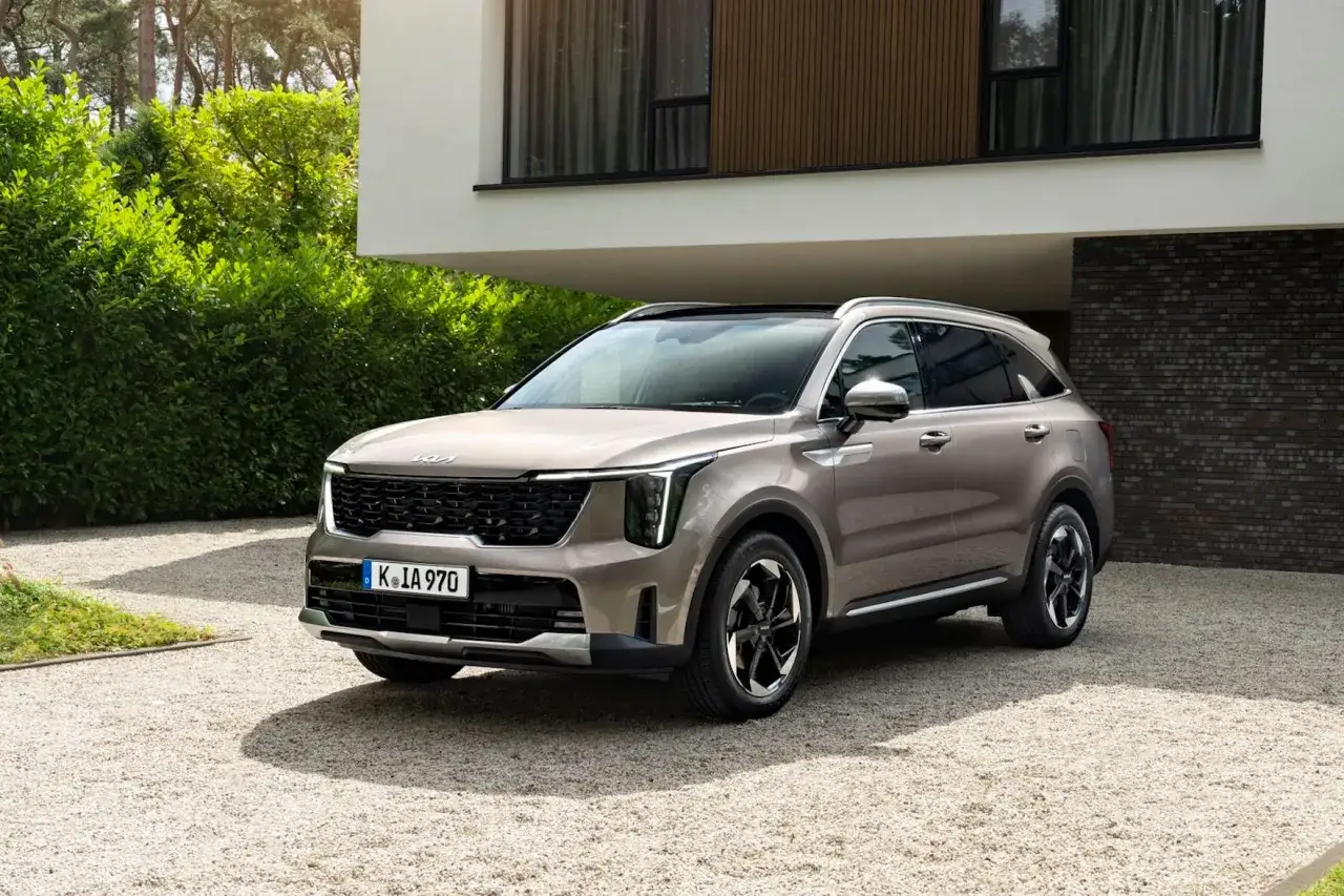 Elegancki, beżowy SUV z rodziny kia modele, zaparkowany przed nowoczesnym domem z ceglaną ścianą i bujną zielenią.