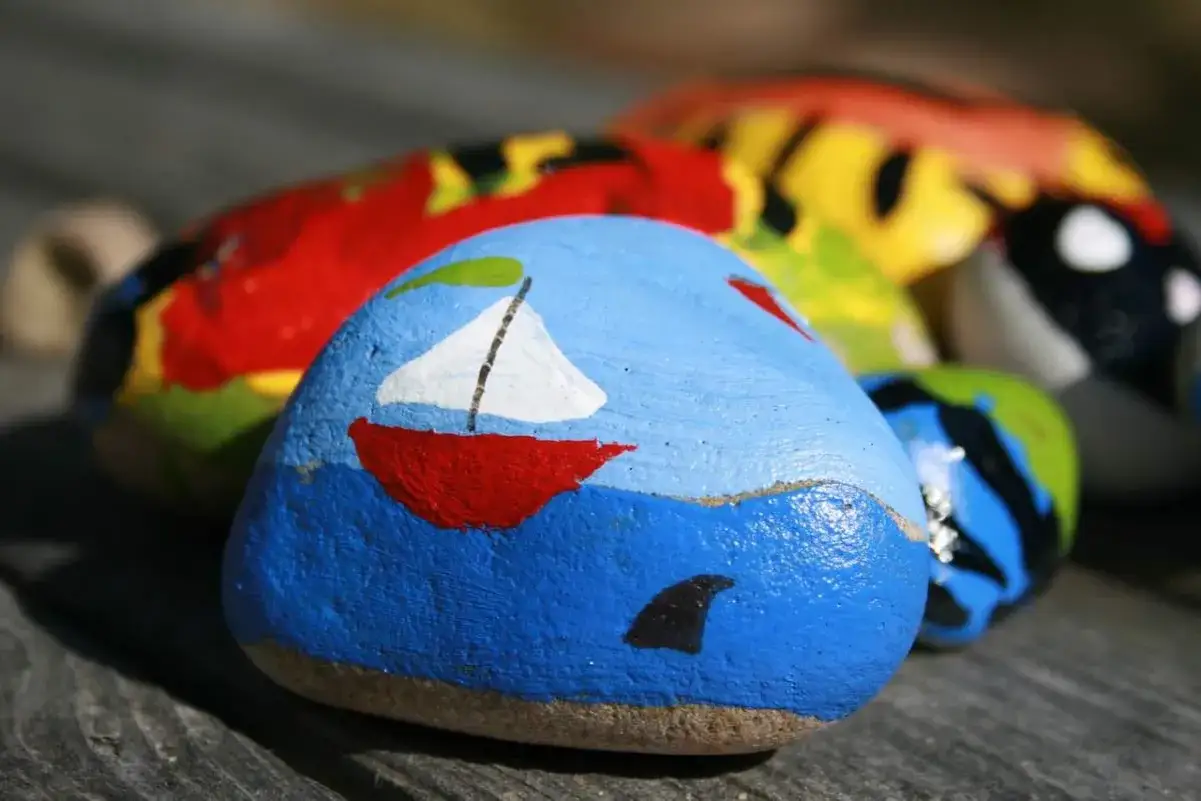 Piedras pintadas con ideas creativas: un barco rojo navega en mar azul con aleta de tibur&oacute;n.