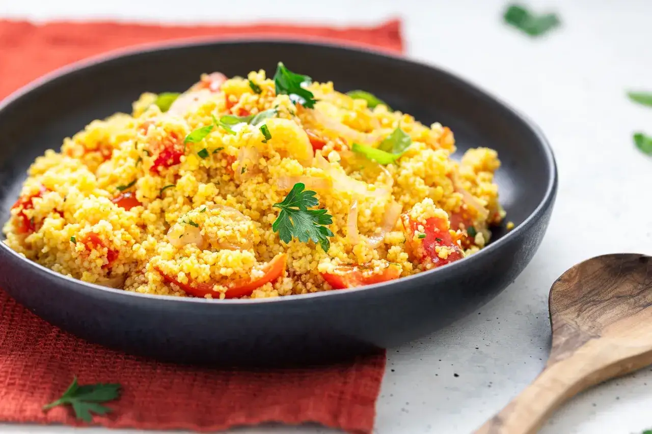 Un'insalata di cous cous colorata con pomodorini, cipolle e prezzemolo fresco, servita in una ciotola scura.