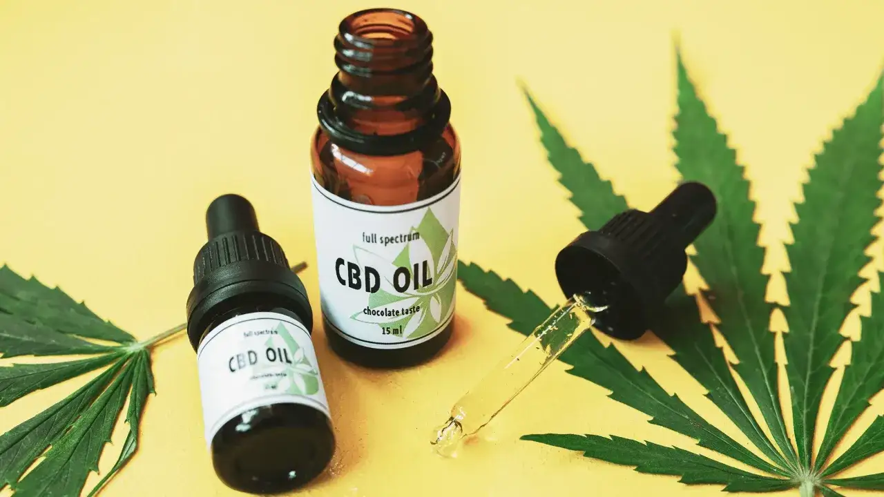 Olej CBD na co? Dwie buteleczki z kroplomierzem i liście konopi na żółtym tle.