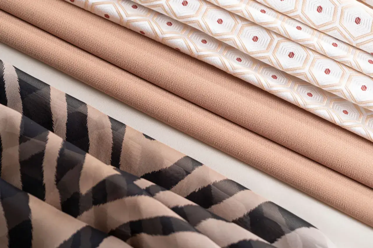 Tessuti piegati: uno con fantasia zebrata, uno liscio color sabbia, uno con rombi beige e puntini rossi.