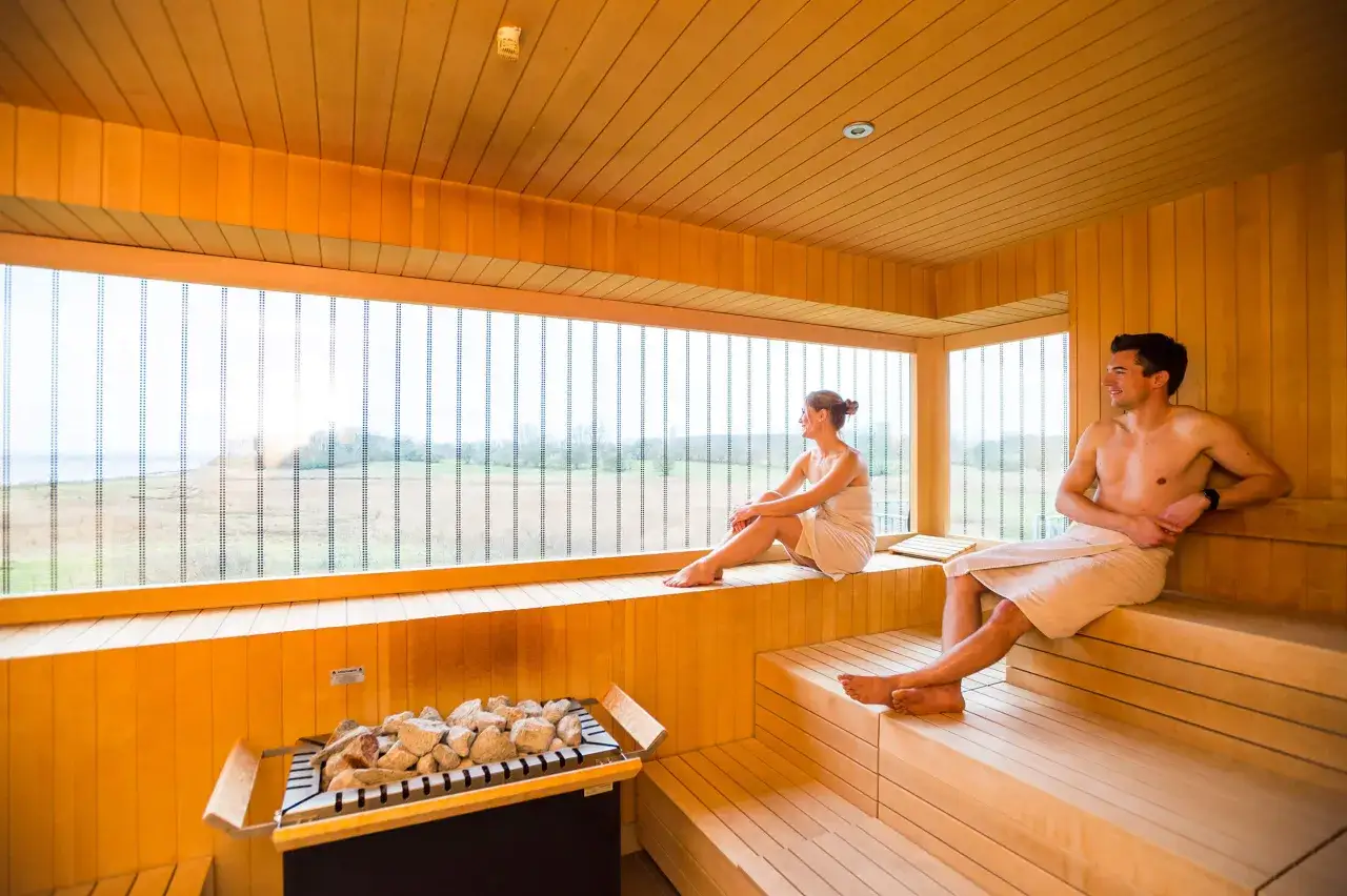 Ein Paar entspannt in einer modernen Therme an der Ostsee. Die holzverkleidete Sauna mit großem Fenster bietet Blick auf die Natur.