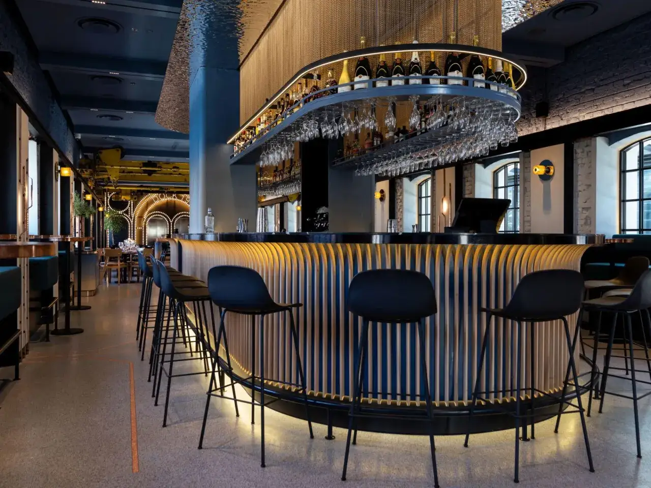 Nowoczesny bar z drewnianą ladą i czarnymi hokerami. Na półkach nad ladą stoją butelki alkoholu, a pod nimi wiszą kieliszki. W tle widać stoliki i krzesła. Mateusz Gessler z pewnością doceniłby ten wystrój.