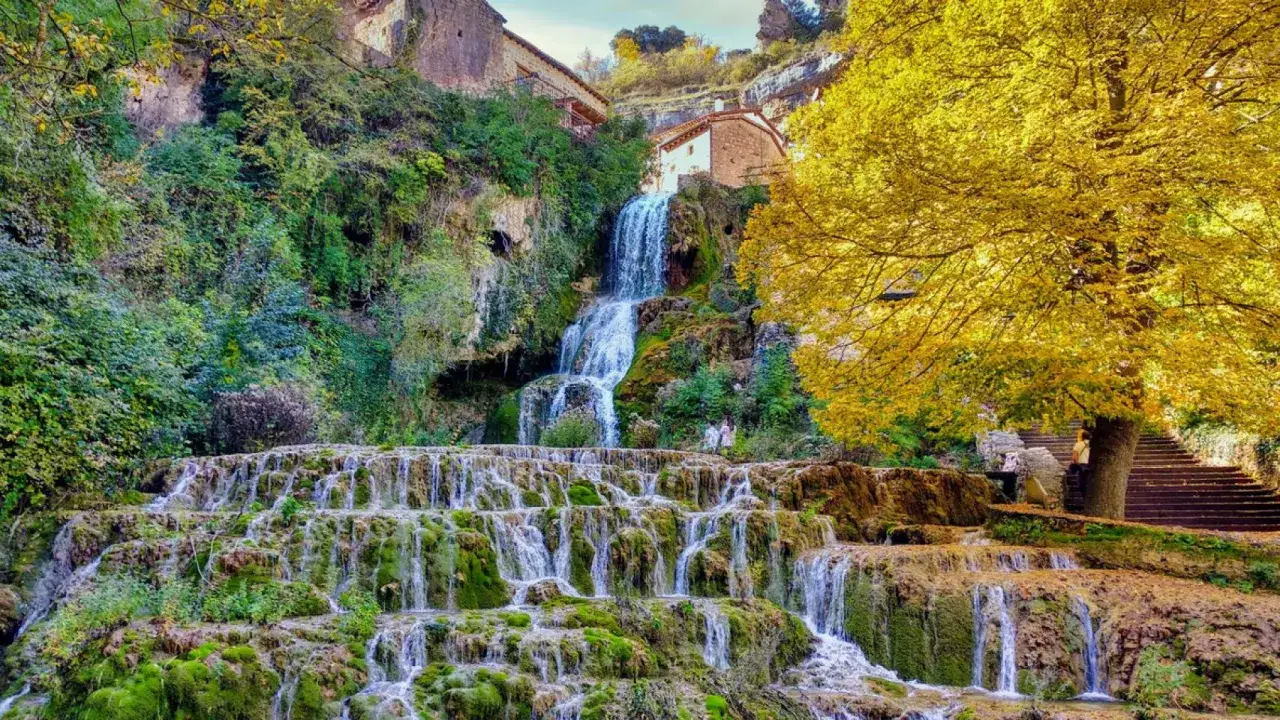 Cascada en Orbaneja del Castillo. Impresionante naturaleza que ver en Burgos, con casas integradas en la roca y un &aacute;rbol de hojas doradas.
