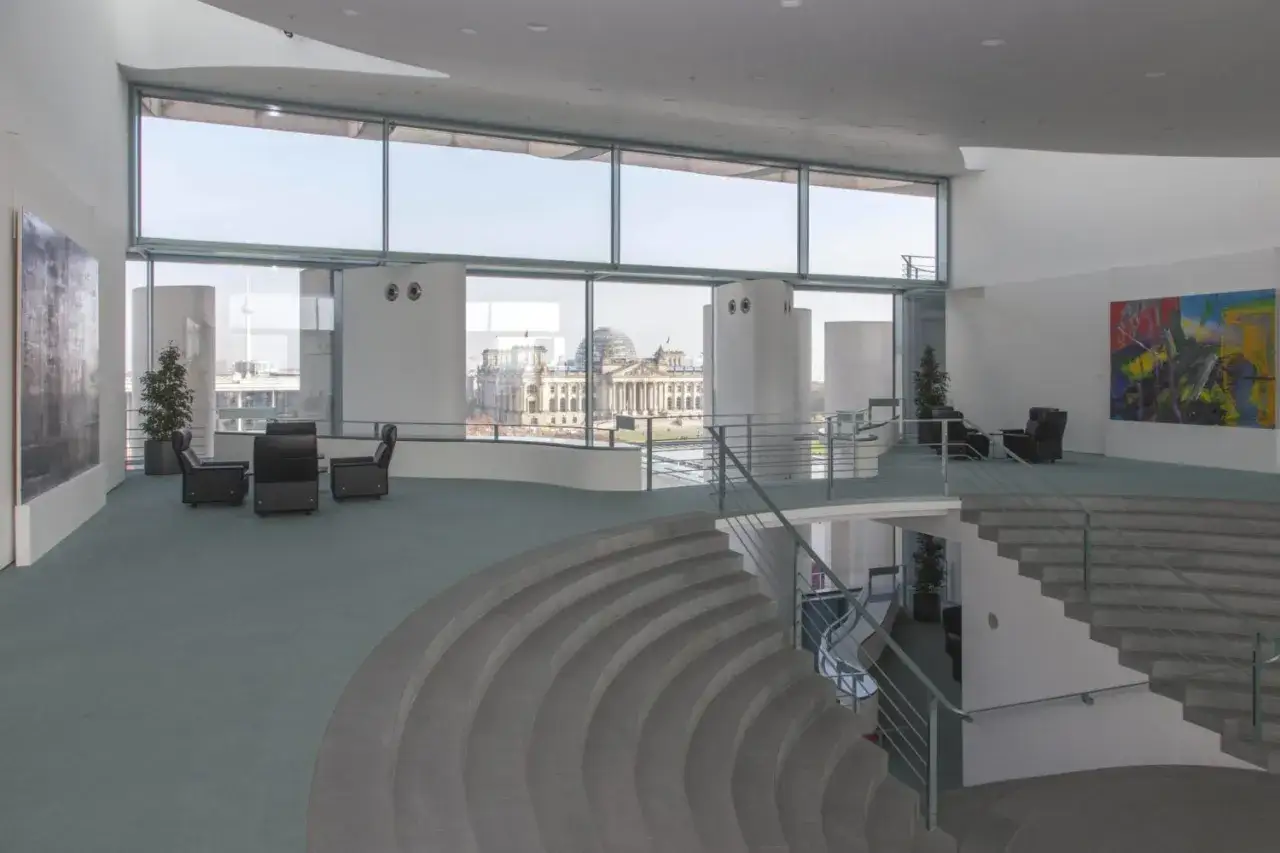 Modernes Foyer im Bundeskanzleramt innen mit Blick auf das Reichstagsgeb&auml;ude. Sitzgruppen und eine geschwungene Treppe pr&auml;gen den Raum.