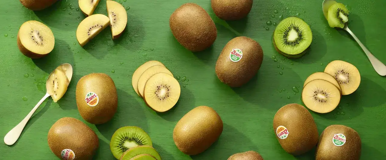 Goldene und grüne Kiwis, einige ganz, andere aufgeschnitten, liegen auf einem grünen Untergrund. Ein Löffel mit Kiwi-Fruchtfleisch. Ein süßer Traum vor dem Schlafen.