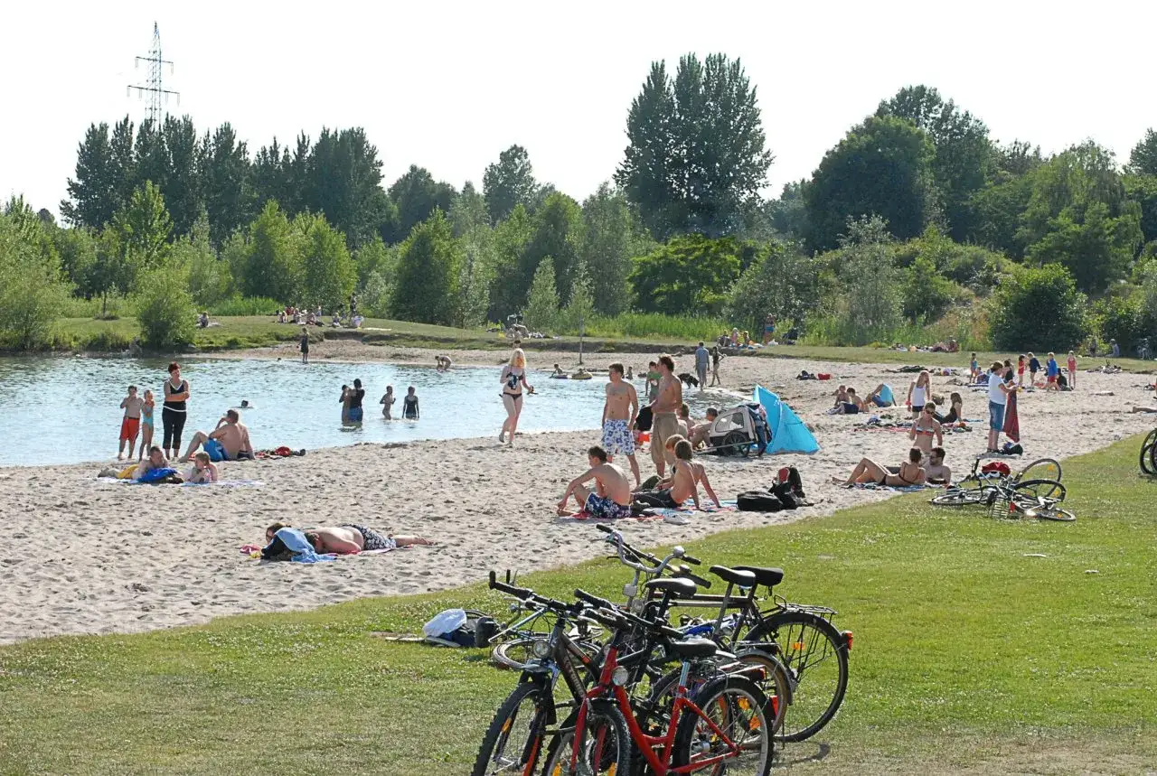 Sonniger Tag am badesee braunschweig. Menschen planschen im Wasser, sonnen sich am Sandstrand und entspannen im Gr&uuml;nen. Fahrr&auml;der stehen bereit.