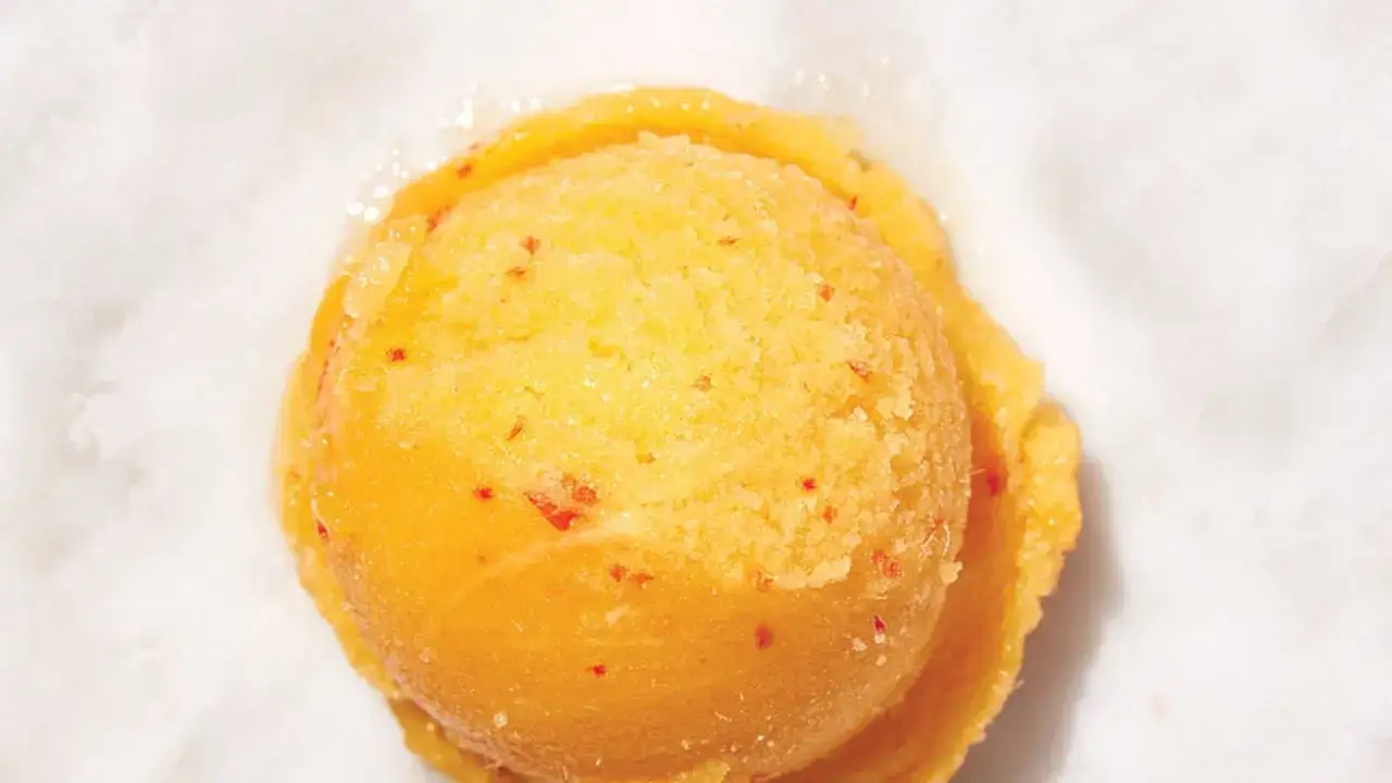 Ein Löffel cremiges Mango-Sorbet mit kleinen roten Fruchtstückchen, das auf weißem Untergrund schmilzt.