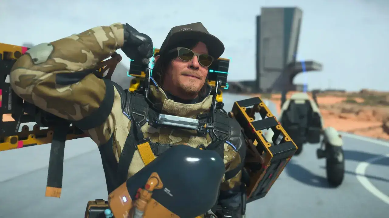 Sam Porter Bridges, con gafas de sol y gorra, listo para una nueva entrega en Death Stranding 2. Lleva su equipo de carga y un veh&iacute;culo de fondo.