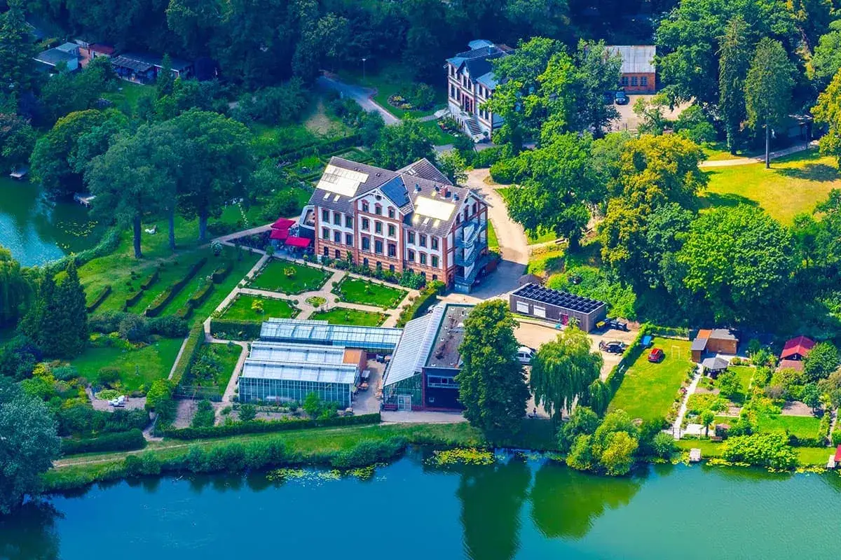 Luftaufnahme des schaugarten am tiefwarensee mit Gewächshäusern, einem Hauptgebäude und einem See.