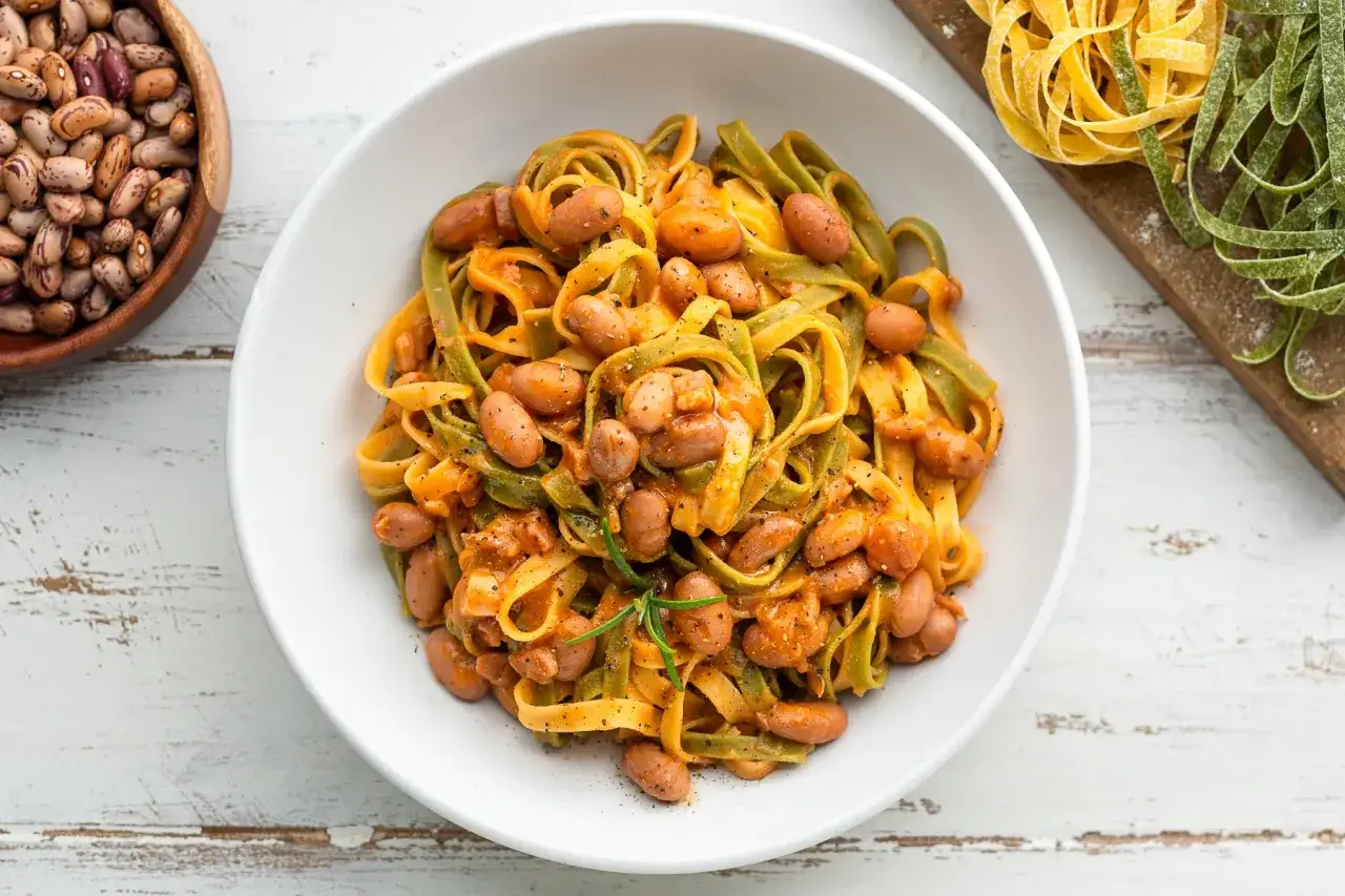 Pasta all'uovo con fagioli borlotti in una ciotola bianca, con fagioli secchi e pasta fresca colorata su un tagliere sullo sfondo.