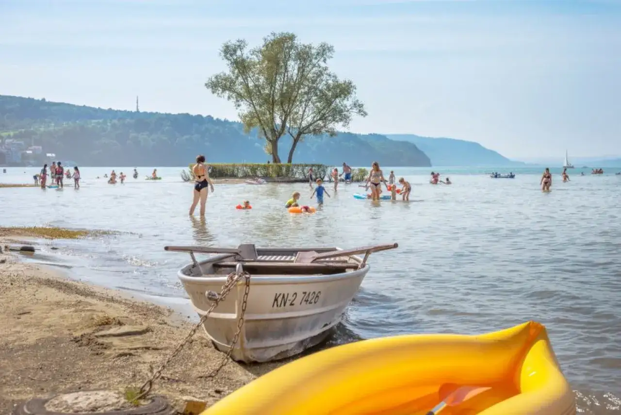 Familien genie&szlig;en einen sonnigen Tag im Strandbad Bodensee. Kinder planschen im Wasser, w&auml;hrend Boote am Ufer liegen.