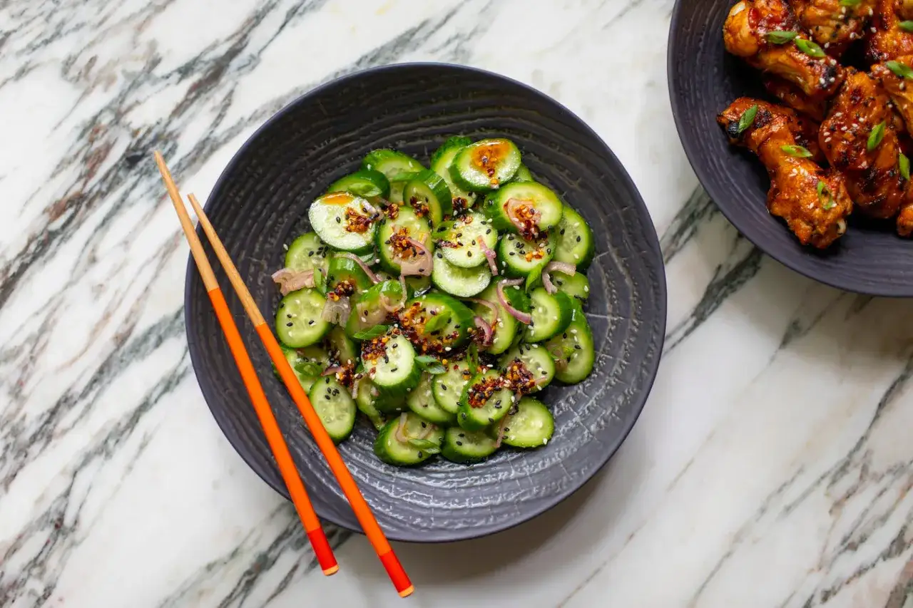 En skål med asiatisk gurksallad, toppad med sesamfrön och chili, bredvid kycklingvingar.